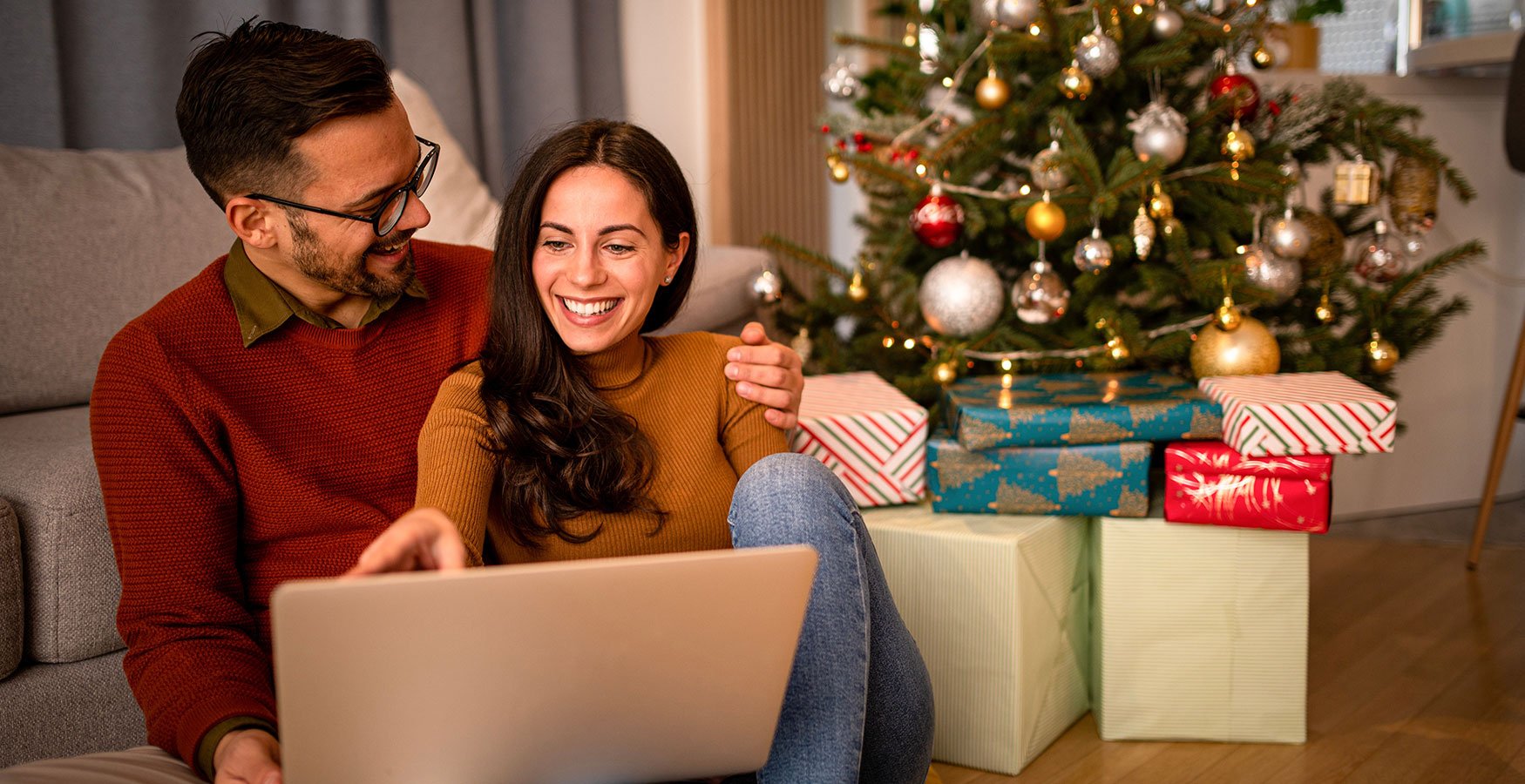 Happy couple smiling while browsing their laptop. Sitting in front of Christmas tree with presents. 