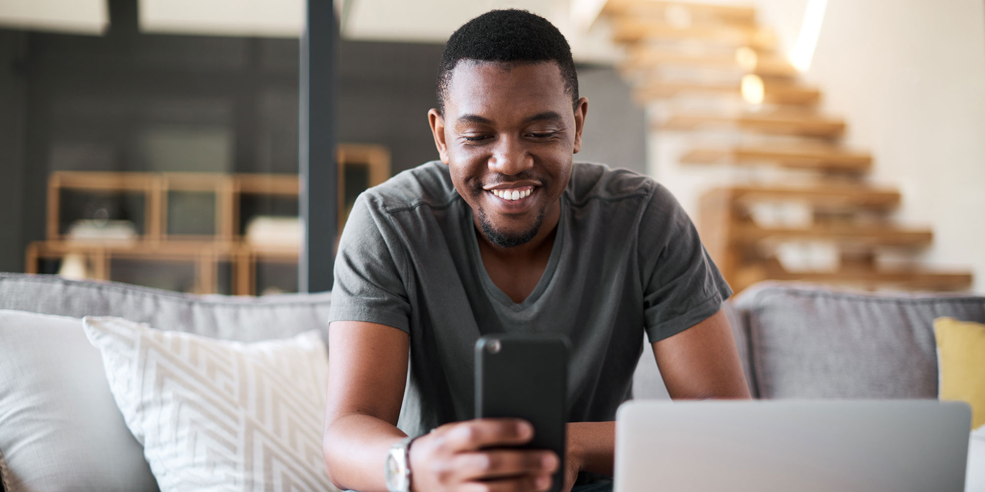 Man smiling while looking at a smartphone, sitting on a couch with a laptop nearby.