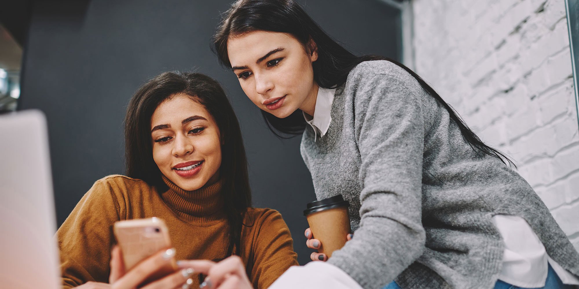 Two women looking at a phone together, one holding a coffee cup, in a modern office setting.